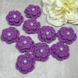 May include: A collection of ten handmade crochet flowers in a vibrant purple hue. Each flower features a central pearl bead, adding a touch of elegance. These decorative floral embellishments are ideal for various craft projects.