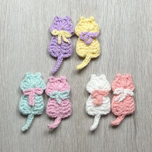 以下が含まれることがあります： ピンク、ブルー、イエロー、パープルなど、さまざまなパステルカラーのクロシェ編み猫のアップリケ6個。各猫の首には小さなリボンが結ばれています。