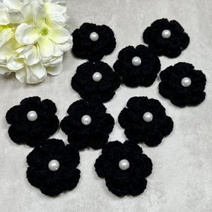May include: A collection of ten black crochet flower appliques, each with a white pearl bead center. The flowers are arranged on a light gray surface, with a white flower in the upper left corner.