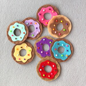 Può includere: Una collezione di ciambelle in feltro fatte a mano in vari colori. Ogni ciambella presenta una base marrone con glassa colorata e piccole perline. Le ciambelle includono glassa rosa, viola, blu, gialla, rossa e marrone.