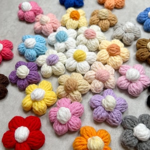 May include: A collection of crocheted flower embellishments in various colors, including pink, yellow, purple, blue, orange, and white. The flowers have a 3D appearance and are made with yarn.