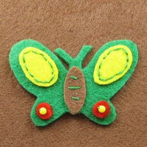 Puede incluir: Una mariposa de fieltro verde con detalles amarillos y acentos rojos y amarillos. La mariposa tiene óvalos amarillos en sus alas, delineados con costuras verdes. El cuerpo es marrón y las alas son verdes. Dos círculos rojos con cuentas amarillas están en la parte inferior.