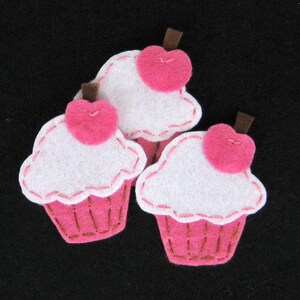 Può includere: Tre decorazioni per cupcake in feltro con glassa rosa e un cuore rosa in cima. I cupcake sono bianchi con cuciture rosa attorno ai bordi.