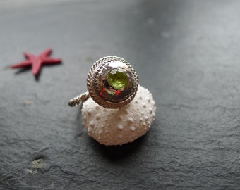 Pode incluir: Um anel de prata com uma gema verde engastada em uma moldura texturizada. O anel é exibido em um ouriço-do-mar branco.