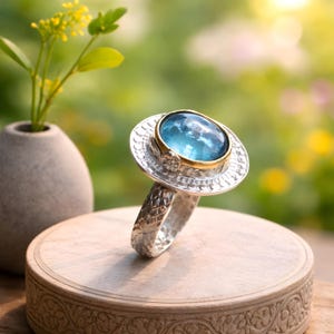 Può includere: Anello d'argento con una grande pietra preziosa ovale blu chiaro incastonata in una lunetta dorata. L'anello ha una fascia strutturata e un'incastonatura decorativa. L'anello è esposto su una superficie di legno.
