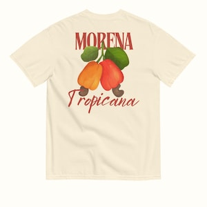 May include: A cream-coloured t-shirt with the word "MORENA" above a graphic of two cashews with green leaves. Below the cashews, the word "Tropicana" is written in a script font. This is a casual wear item.