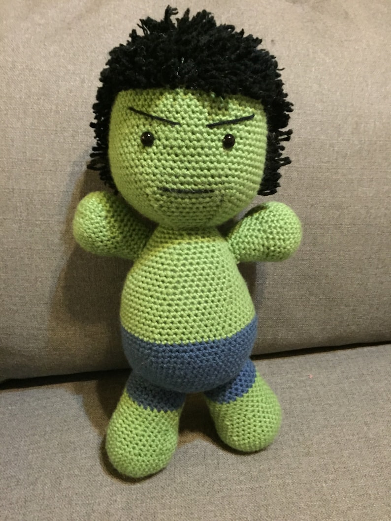 May include: Green crocheted plush toy with black hair and a blue waistband. The toy has a cartoon-like face with black eyes and a frown.
