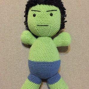 May include: Green crocheted plush toy with a black hairpiece and a blue waistband. The toy has a serious expression with black eyes and a furrowed brow.