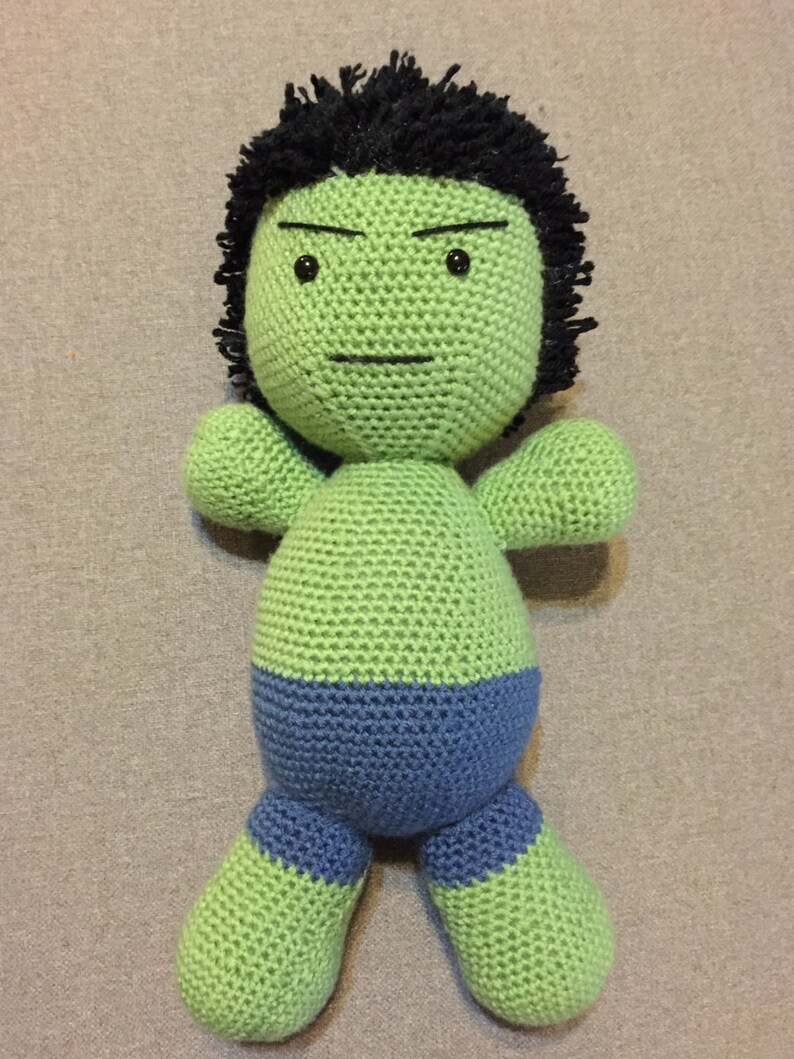 May include: Green crocheted plush toy with a black hairpiece and a blue waistband. The toy has a serious expression with black eyes and a furrowed brow.