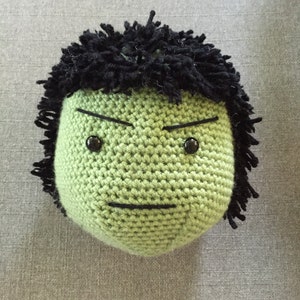 May include: Green crocheted plush toy with a black yarn hair and a frowning face. The toy has black eyes and a black stitched mouth.