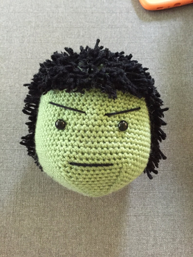 May include: Green crocheted plush toy with a black yarn hair and a frowning face. The toy has black eyes and a black stitched mouth.