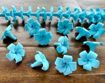 May include: A collection of light blue, flower-shaped decorations. Some are individual blossoms, while others are connected in a chain. The petals are curved and detailed, and the overall effect is delicate and ornamental.