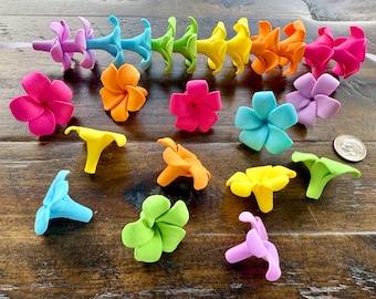 May include: A collection of colorful, handcrafted flower-shaped hair accessories in various shades of pink, blue, green, yellow, orange, and purple. The accessories are arranged on a wooden surface, showcasing their vibrant colors and intricate designs.