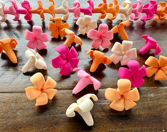 May include: A collection of colorful, handcrafted flower-shaped decorations in shades of pink, orange, and white. Some are arranged in strands, while others are scattered on a wooden surface. These decorative flowers are ideal for crafts.