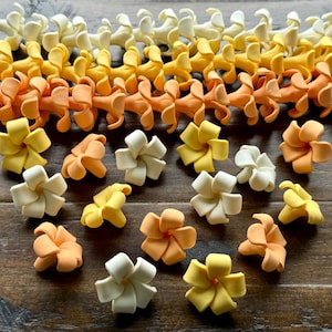 May include: An assortment of handcrafted sugar flowers in various colors, including white, yellow, and orange. Some flowers are arranged in a garland, while others are scattered on a wooden surface. These edible decorations are perfect for cakes and cupcakes.
