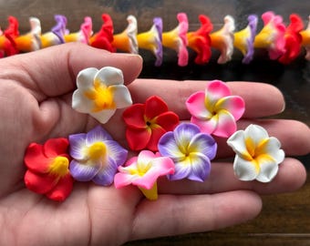 May include: A hand holds several colorful plumeria flower charms in shades of red, white, pink, and purple with yellow centers. A row of similar charms is in the background. These charms are ideal for jewelry making or craft projects.
