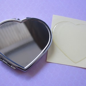 May include: A silver heart-shaped compact mirror with a smooth, reflective surface. The mirror is closed and sitting on a light purple surface next to a white heart-shaped sticker.