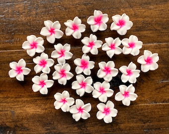 May include: A collection of white plumeria flowers with bright pink centers. The flowers are arranged on a dark brown wooden surface. These decorative flowers are ideal for crafting or adding a tropical touch to any project.