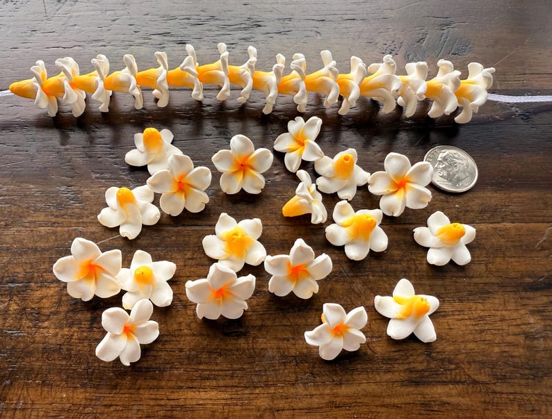 May include: A collection of white and yellow artificial flowers with orange centers. Some are arranged in a chain, while others are scattered. A US quarter is included for size reference. The flowers are on a dark wooden surface.