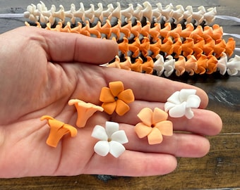 May include: A hand holds several small, decorative items. There are orange and white flower-shaped decorations, along with orange mushroom-shaped decorations. In the background are rows of orange and white decorative pieces.