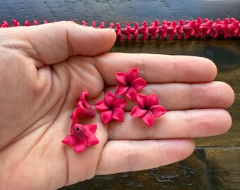 May include: Close-up of several small, vibrant pink flower-shaped beads held in a hand. The flowers have five petals and a dark center. A string of similar beads is visible in the background.