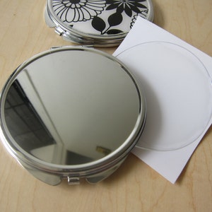 May include: A silver compact mirror with a round, reflective surface. The mirror is closed and has a black and white floral design on the outside.