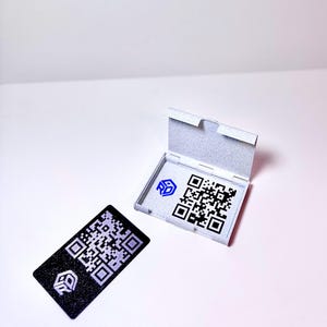 Könnte beinhalten: Eine schwarz-weiße QR-Code-Karte mit einem Logo, neben einer offenen, hellgrauen Box mit einem QR-Code und Logo. Die Karte ist rechteckig und hat eine strukturierte Oberfläche. Die Box ist geöffnet und zeigt den QR-Code.
