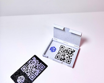 Puede incluir: Una tarjeta de código QR en blanco y negro con un logotipo, junto a una caja gris claro abierta con un código QR y un logotipo. La tarjeta es rectangular y tiene una superficie texturizada. La caja está abierta, revelando el código QR.