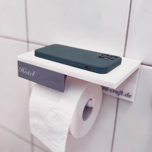 May include: A white wall-mounted toilet paper holder with a shelf, holding a roll of toilet paper and a teal phone case. The word "Hotel" is printed on the holder. The holder is mounted on white tiled wall.