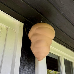 Op de afbeelding: Een terracottakleurig keramisch bijenhuis, gevormd als een afgeronde kegel, hangt aan de dakgoot van een huis.