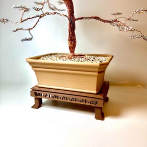 Könnte beinhalten: Eine handgefertigte Bonsai-Baumskulptur mit einem Stamm und Ästen aus Kupferdraht sowie Blättern aus Silberdraht. Der Baum steht in einem beigen rechteckigen Topf, der mit kleinen Steinen gefüllt ist, auf einem dunkelbraunen Holzständer. Der Bonsai ist ein dekoratives Kunstwerk.