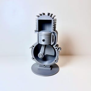 Könnte beinhalten: Graues 3D-gedrucktes Modell der inneren Komponenten eines Motors. Der Querschnitt zeigt Kolben, Zylinder und Kurbelwelle. Das Modell ist auf einem kreisförmigen Sockel montiert, um die Funktionsweise des Motors zu veranschaulichen.