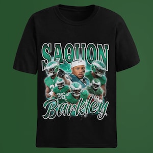 May include: Black t-shirt featuring a graphic of Saquon Barkley in a Philadelphia Eagles uniform. The design includes the name "Saquon Barkley" in a green, vintage-style font, with images of the player in action.