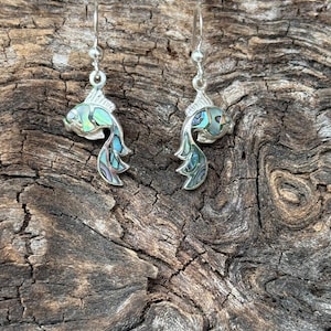 May include: A pair of silver fish earrings with iridescent abalone shell accents. The fish have detailed fins and tails, and are suspended from simple silver hooks. The earrings are displayed against a textured, dark wood background.