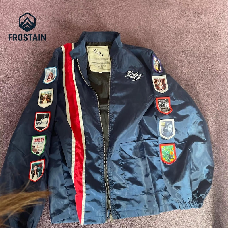 Puede incluir: Chaqueta bomber azul marino con una franja roja y blanca en el lado izquierdo. La chaqueta presenta m&uacute;ltiples parches con im&aacute;genes y el texto "LDR" en el pecho y la espalda. El logotipo de la marca "FROSTAIN" est&aacute; en la esquina superior izquierda.