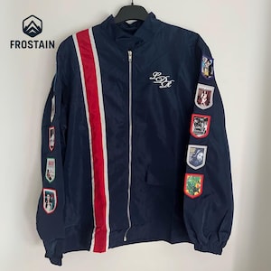 May include: A navy blue zip-up jacket with a red and white stripe down the front. The jacket has embroidered patches on the sleeves and the initials "LDR" in white script. The jacket is hanging on a black hanger.