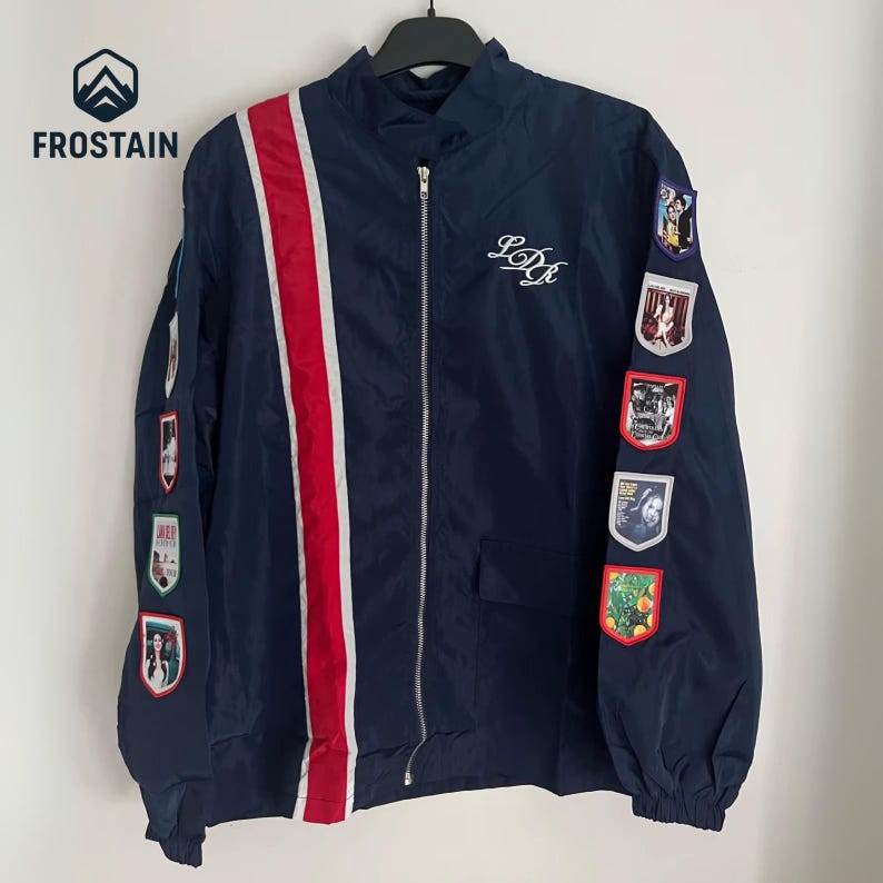 Puede incluir: Chaqueta azul marino con cremallera y una franja roja y blanca en la parte delantera. La chaqueta presenta parches bordados en las mangas y las iniciales "LDR" en escritura blanca. La chaqueta est&aacute; colgada de una percha negra.