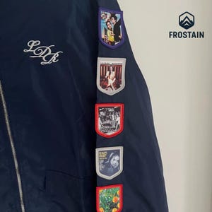 Puede incluir: Chaqueta azul marino con cierre de cremallera y letras blancas cursivas "LDR". La chaqueta presenta varios parches coloridos con im&aacute;genes. La marca "FROSTAIN" est&aacute; impresa en la parte superior derecha.