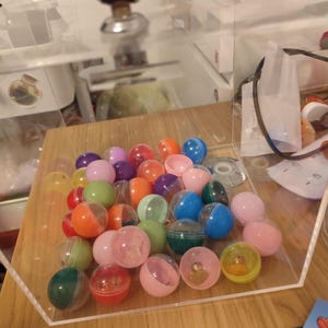 Op de afbeelding: Een doorzichtige acryl displaybox met een zilveren slot en sleutel aan de bovenkant, gevuld met een verzameling kleine, ronde, veelkleurige plastic capsules in roze, blauw, groen, oranje en paars, rustend op een houten oppervlak met liniaal.