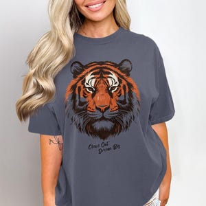Könnte beinhalten: Ein schieferblaues T-Shirt mit einem Tigerkopf-Motiv in Orange, Braun und Weiß. Der Text "Close Out Dream Big" ist darunter aufgedruckt. Das T-Shirt ist ein lässiger Stil, geeignet für den täglichen Gebrauch.