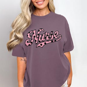 Könnte beinhalten: Ein mauvefarbenes T-Shirt mit dem Wort "Nurse" in einem rosa Retro-Schriftzug mit schwarzen Punkten. Das Shirt hat kurze Ärmel und eine lockere Passform.