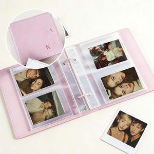 Op de afbeelding: Een lichtroze fotoalbum met de initialen "A" en "K" en een bloemmotief op de kaft. Het album is open en toont verschillende foto's in transparante plastic hoezen. Er ligt ook een losse foto op het oppervlak.