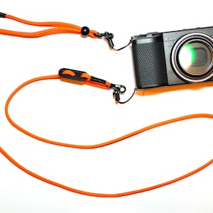 May include: A black digital camera with a bright orange neck strap. The camera lens is centered, with the letters "GR" visible on the right side. The strap has black connectors and a black adjustment bead.