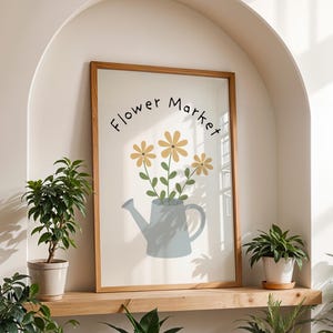 May include: A framed print with a wooden frame featuring a floral illustration. The artwork depicts yellow flowers in a blue watering can, with the words "Flower Market" in a handwritten font. The print is displayed on a wooden shelf with potted plants.