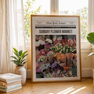 May include: A framed print of a newspaper front page titled "Sunday Flower Market" with a vibrant floral arrangement. The print is in a light wood frame, set against a window with sheer curtains, and surrounded by potted plants and a stack of books.