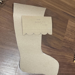 May include: A tan cardboard template for a Christmas stocking with a separate rectangular piece labeled 'Xmas stocking trim x1 (felt)' featuring a scalloped bottom edge. The main piece is shaped like a stocking and both are made of stiff paper.