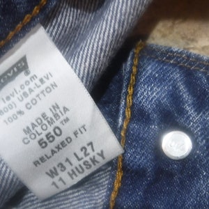 May include: A white Levi's tag with black text, showing the brand name, website, phone number, and fabric content. The tag also includes the size and fit information: 550 Relaxed Fit, W31 L27, 11 Husky. The tag is attached to a pair of blue denim jeans.