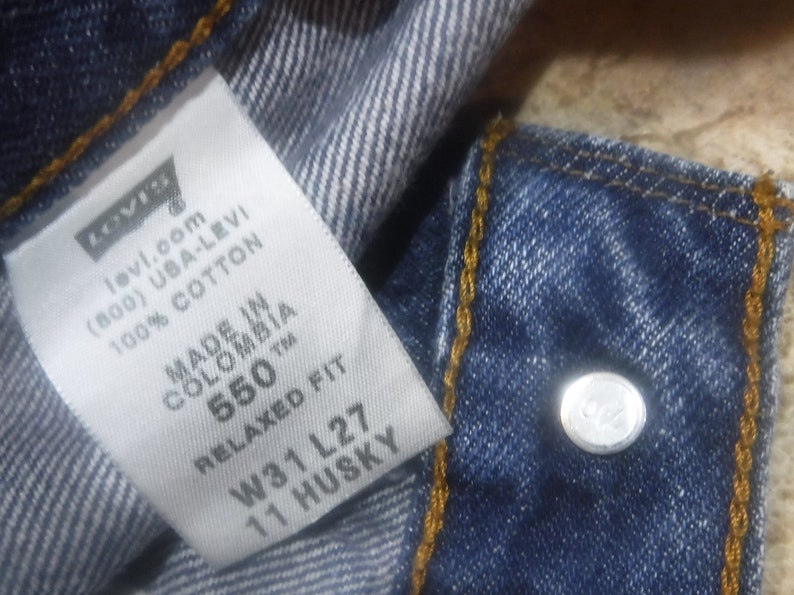 May include: A white Levi's tag with black text, showing the brand name, website, phone number, and fabric content. The tag also includes the size and fit information: 550 Relaxed Fit, W31 L27, 11 Husky. The tag is attached to a pair of blue denim jeans.