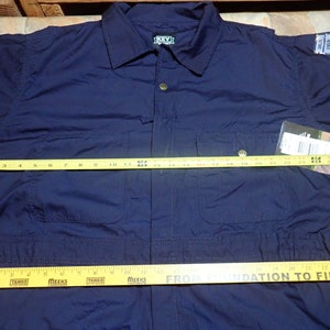 May include: A navy blue workwear jumpsuit with long sleeves and a button-up front. The jumpsuit has two chest pockets and two front pockets. A yellow measuring tape is placed across the chest of the jumpsuit, measuring 28 inches.