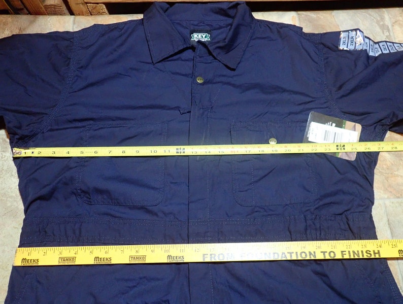 May include: A navy blue workwear jumpsuit with long sleeves and a button-up front. The jumpsuit has two chest pockets and two front pockets. A yellow measuring tape is placed across the chest of the jumpsuit, measuring 28 inches.